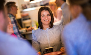 Bli en av oss huvudbild där Amelie Johansson sitter vid ett runt bord med andra anställda på Baker Tilly Halmstad