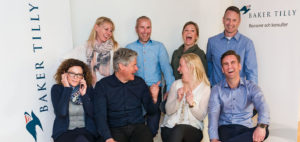 Galleri huvudbild där Charlotte Falk, Jan Carlsson, Jessica Herrlin, Linda Sturesson, Fredrik Wihed och Henrik Andersson skrattar och är glada vid en skylt med Baker Tilly Revisorer och Konsulter skrivet